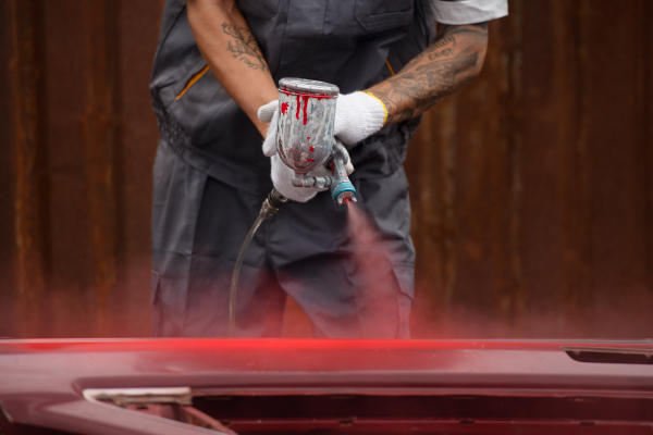 Un trabajador con guantes blancos usa una pistola para aplicar pintura roja en una superficie. La imagen muestra un primer plano de la pistola de pintura en acción, mientras el trabajador sostiene firmemente la herramienta, sugiriendo un proceso de pintura industrial o automotriz.