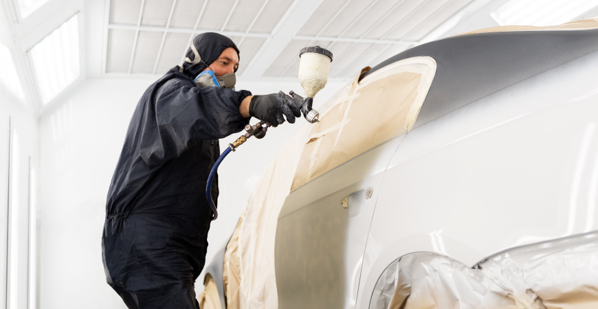 Un hombre en un taller de pintura automotriz, aplicando pintura a un automóvil con una pistola de pulverización. La persona lleva un traje de protección y guantes, y el automóvil está parcialmente cubierto con papel para proteger las áreas que no se van a pintar. Esta escena ilustra el proceso de pintura automotriz, esencial para el mantenimiento y personalización de vehículos.