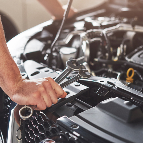 La imagen muestra a una persona sosteniendo dos llaves inglesas mientras trabaja en el motor de un automóvil. La persona está enfocada en la parte delantera del vehículo, con el capó levantado, lo que sugiere que está realizando tareas de mantenimiento o reparación.