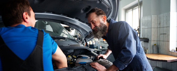 Un mecánico está trabajando en el motor de un automóvil. La imagen muestra al mecánico inclinado sobre el motor, utilizando herramientas para realizar reparaciones o mantenimiento. El entorno parece ser un taller mecánico, con varias herramientas y equipos visibles en el fondo. La escena ilustra el trabajo manual y técnico necesario para mantener los vehículos en buen estado de funcionamiento.