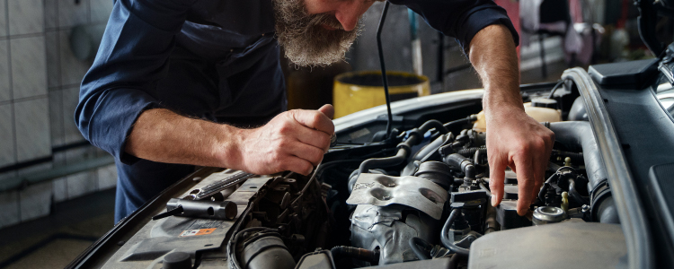 Un mecánico está trabajando en el motor de un automóvil. La imagen muestra al mecánico inclinado sobre el motor, utilizando herramientas para realizar reparaciones o mantenimiento. El entorno parece ser un taller mecánico, con varias herramientas y equipos visibles en el fondo. La escena ilustra el trabajo manual y técnico necesario para mantener los vehículos en buen estado de funcionamiento.