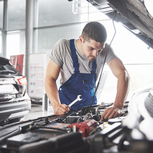 Un mecánico, vestido con un mono de trabajo azul y una camisa gris, revisa el motor de un automóvil en un taller. Sostiene una llave inglesa y parece concentrado en su tarea. Al fondo, se observa otro vehículo, indicando que es un espacio activo de reparación y mantenimiento de coches. La imagen subraya la importancia del trabajo mecánico para garantizar el buen funcionamiento de los vehículos.