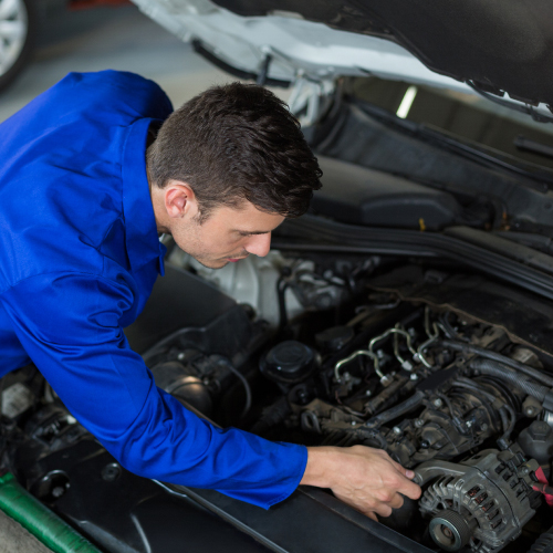 Mecánico con una bata azul trabajando en el motor de un automóvil. La persona está inclinada sobre el motor, aparentemente ajustando o reparando una parte del mismo. El capó del automóvil está levantado, permitiendo una vista clara del motor y de las manos de la persona que están manipulando una herramienta. Es una escena típica de mantenimiento y reparación vehicular, destacando la destreza y precisión necesarias para mantener los coches en buen estado.