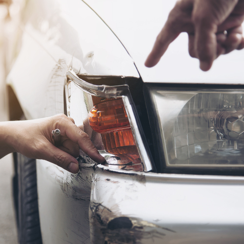 La imagen muestra un primer plano de la parte delantera de un automóvil que ha sufrido daños. Se pueden ver dos manos señalando y tocando la zona dañada, que incluye un faro roto y abolladuras en la carrocería. La imagen ilustra un accidente automovilístico y la inspección de los daños resultantes.