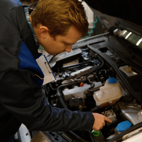 Un mecánico está inspeccionando el motor de un automóvil. El mecánico está señalando una parte específica del motor mientras sostiene una carpeta con documentos. La imagen muestra el motor del automóvil con varios componentes visibles, incluyendo un depósito de líquido refrigerante y otros elementos del sistema del motor. Esta imagen es relevante porque ilustra una situación común en la que un mecánico realiza una revisión o mantenimiento de un vehículo, lo cual es esencial para el buen funcionamiento y la seguridad del automóvil.