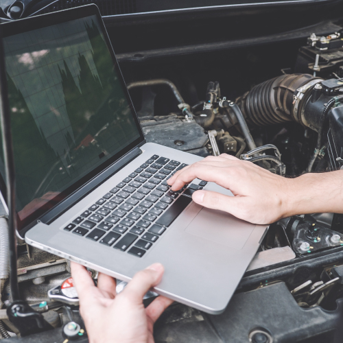 Una persona está utilizando una computadora portátil mientras trabaja en el motor de un automóvil. La computadora está apoyada sobre el motor, y las manos de la persona están sobre el teclado, posiblemente diagnosticando o reparando el vehículo con la ayuda de software especializado.