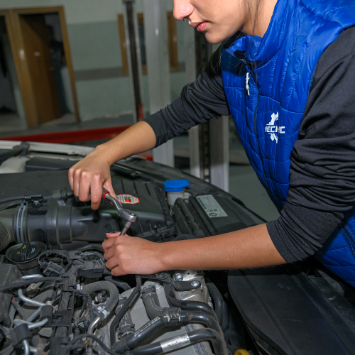 La imagen muestra a una persona con un chaleco azul y una camiseta negra trabajando en el motor de un automóvil. La persona está usando una herramienta para ajustar o reparar una parte del motor.