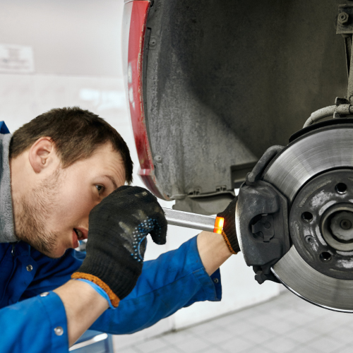 Un hombre con traje azul trabajando en el sistema de frenos de un automóvil. La persona está utilizando una herramienta para ajustar o reparar el freno de disco. La imagen es relevante porque ilustra una tarea común en el mantenimiento de vehículos, que es esencial para la seguridad en la conducción.