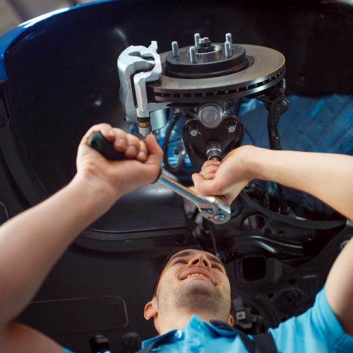 La imagen muestra a una persona trabajando en la reparación o mantenimiento de un vehículo. Utiliza una llave de trinquete para ajustar o aflojar una pieza en el sistema de suspensión o frenos del automóvil. Se puede ver un disco de freno y otras partes mecánicas del vehículo. La imagen destaca el trabajo técnico y especializado que se realiza en el mantenimiento de vehículos, subrayando la importancia de las habilidades mecánicas y el uso de herramientas adecuadas.