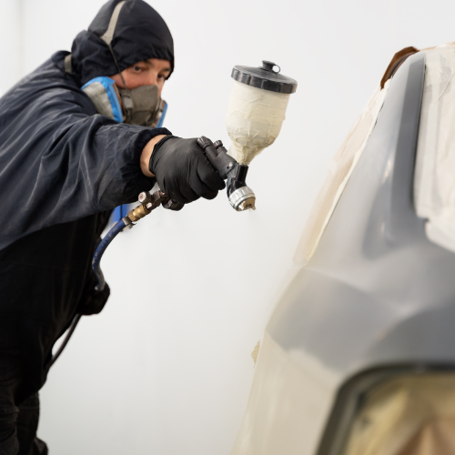 Un trabajador con equipo de protección, incluyendo guantes y una máscara, está pintando un vehículo con una pistola de pintura en un taller. La parte del vehículo que se está pintando está parcialmente cubierta con cinta adhesiva y papel para proteger las áreas que no deben ser pintadas.