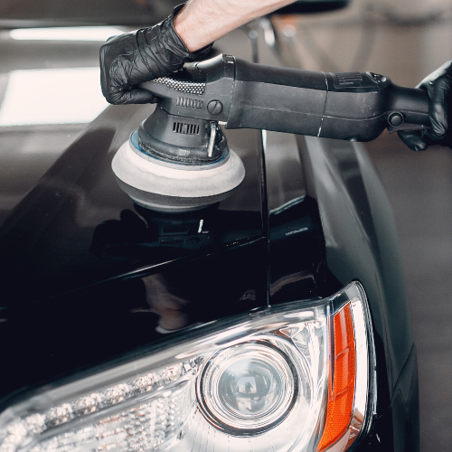 Un trabajador con guantes negros está utilizando una pulidora eléctrica para pulir la superficie de un automóvil negro. La imagen muestra un primer plano del faro delantero del automóvil y la pulidora en acción, destacando el proceso de detallado y mantenimiento del vehículo.