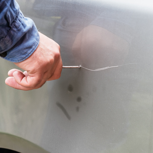 Una mano sosteniendo una herramienta metálica está rayando la superficie de un automóvil plateado.