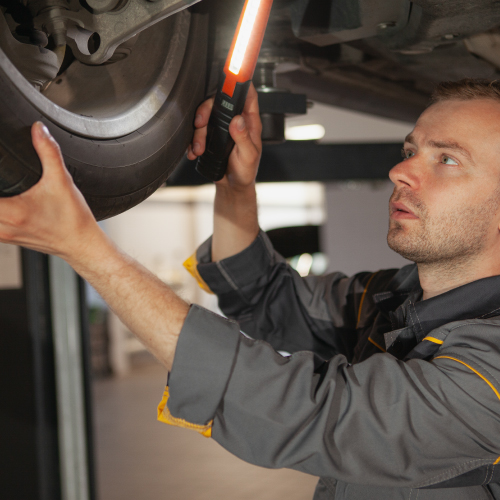 Un mecánico inspeccionando la parte inferior de un vehículo levantado. Está sosteniendo una luz de trabajo con una mano y con la otra mano está tocando una rueda. El mecánico lleva un uniforme de trabajo gris con detalles amarillos.