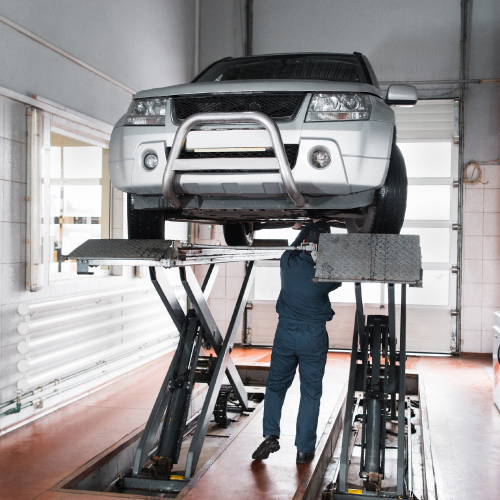 Un mecánico trabaja bajo un coche elevado en un taller, utilizando una plataforma hidráulica para acceder a la parte inferior del vehículo.