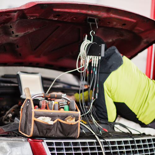 Un mecánico está trabajando en el motor de un automóvil con el capó levantado. Lleva una chaqueta de color verde y negro y está utilizando varios cables conectados a un dispositivo de diagnóstico. En primer plano, hay una caja de herramientas con varias herramientas visibles, como destornilladores y llaves. La imagen muestra un entorno de taller mecánico, destacando la importancia del mantenimiento y diagnóstico de vehículos para asegurar su correcto funcionamiento.