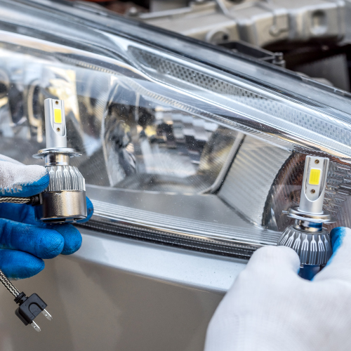 Dos manos con guantes sostienen bombillas LED frente al faro de un automóvil. Las bombillas presentan un diseño metálico con aletas de disipación de calor y un conector eléctrico, sugiriendo que se están instalando o reemplazando en el faro del vehículo.