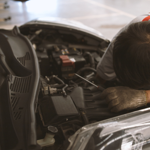 Un mecánico está trabajando en el motor de un automóvil. Se puede ver la parte delantera del vehículo con el capó levantado, y el mecánico está inclinado sobre el motor, usando guantes de trabajo. La imagen muestra una escena típica de mantenimiento o reparación de un automóvil, lo cual es relevante para ilustrar trabajos de mecánica automotriz.