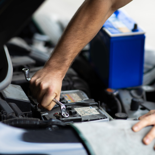 Un mecánico está utilizando una llave para ajustar o aflojar una conexión en la batería de un automóvil. En el fondo, se puede ver una batería de repuesto de color azul. La imagen muestra una actividad común de mantenimiento de vehículos, que es relevante para aquellos interesados en la mecánica automotriz o que necesitan realizar reparaciones en sus propios vehículos.