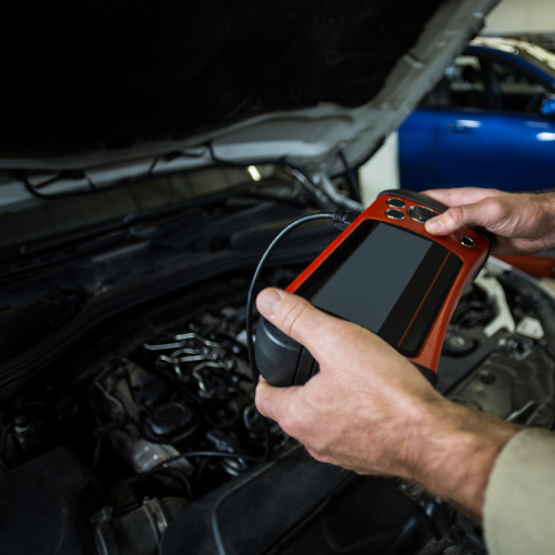 Un mecánico está utilizando una herramienta de diagnóstico automotriz para revisar el motor de un vehículo. La herramienta es de color rojo y negro y está conectada al motor mediante un cable. El capó del vehículo está abierto, permitiendo el acceso al motor.