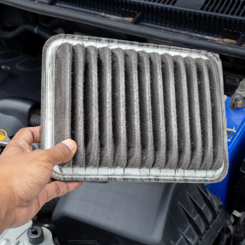 Una mano sostiene un filtro de aire de automóvil sucio, con pliegues visibles llenos de polvo y suciedad. El filtro está ubicado frente al compartimento del motor de un vehículo, donde se pueden ver partes del motor y una batería azul. La imagen es relevante porque muestra la importancia del mantenimiento del vehículo, específicamente la necesidad de cambiar el filtro de aire para asegurar un rendimiento óptimo del motor y una mejor eficiencia de combustible.