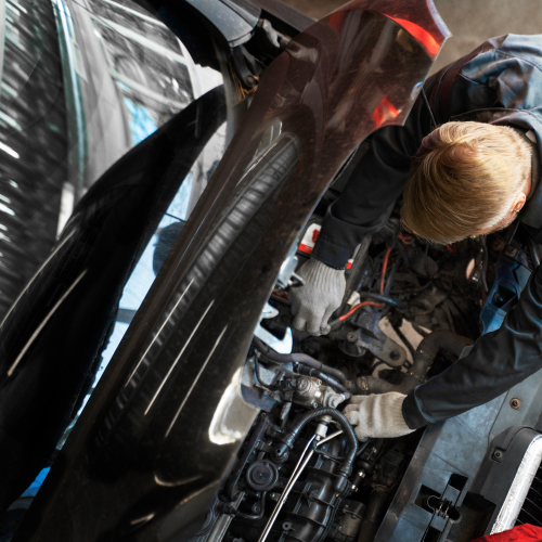 Un mecánico está trabajando en el motor de un automóvil. La imagen muestra una vista desde arriba del mecánico, que lleva guantes y está inclinado sobre el motor, realizando reparaciones o mantenimiento. La escena destaca la importancia del trabajo técnico y especializado en el mantenimiento automotriz.