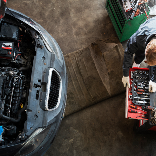 Un mecánico está trabajando en un automóvil en un taller. El capó del automóvil está abierto, mostrando el motor y otros componentes internos. A la derecha, el mecánico está organizando o seleccionando herramientas de una caja de herramientas roja. Hay una alfombra o tapete en el suelo, probablemente para que el mecánico se arrodille o se acueste mientras trabaja. En el fondo, se puede ver un carrito verde con más herramientas y equipos.