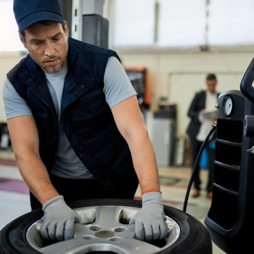 Un mecánico está trabajando en un taller de automóviles. Lleva guantes y está manipulando una llanta de automóvil. En el fondo, se puede ver a otra persona de pie, posiblemente supervisando o esperando. La imagen es relevante porque muestra una actividad común en el mantenimiento de vehículos, destacando la importancia del trabajo manual y la atención al detalle en la industria automotriz.