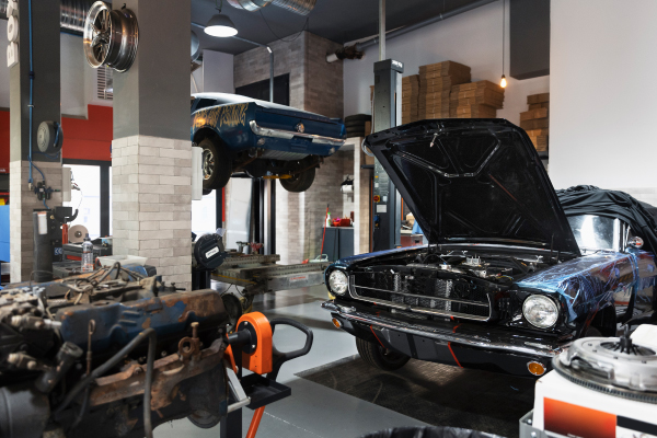 Un taller mecánico con dos autos clásicos: uno elevado en un elevador hidráulico y otro en el suelo con el capó abierto. En primer plano, hay un motor fuera de un vehículo, posiblemente en proceso de reparación o restauración. El taller está organizado, con herramientas y equipos visibles, y cajas apiladas en el fondo. La imagen muestra un entorno de trabajo especializado en la reparación y restauración de autos clásicos, lo que puede ser relevante para los entusiastas de los automóviles y la mecánica.