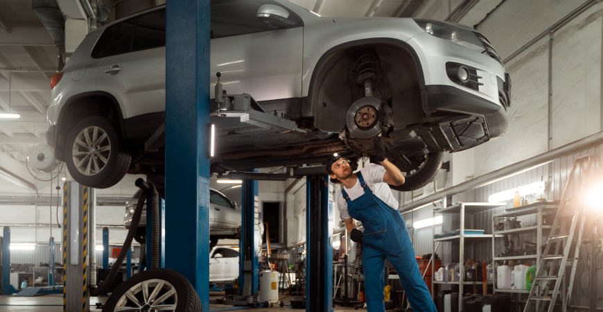 Un automóvil elevado en un taller mecánico, donde un mecánico con un overol azul trabaja en la rueda delantera derecha desmontada. El taller está bien iluminado y equipado con varias herramientas y equipos, incluyendo otro elevador en el fondo y estantes con suministros a la derecha. La imagen resalta un entorno típico de reparación y mantenimiento de vehículos.