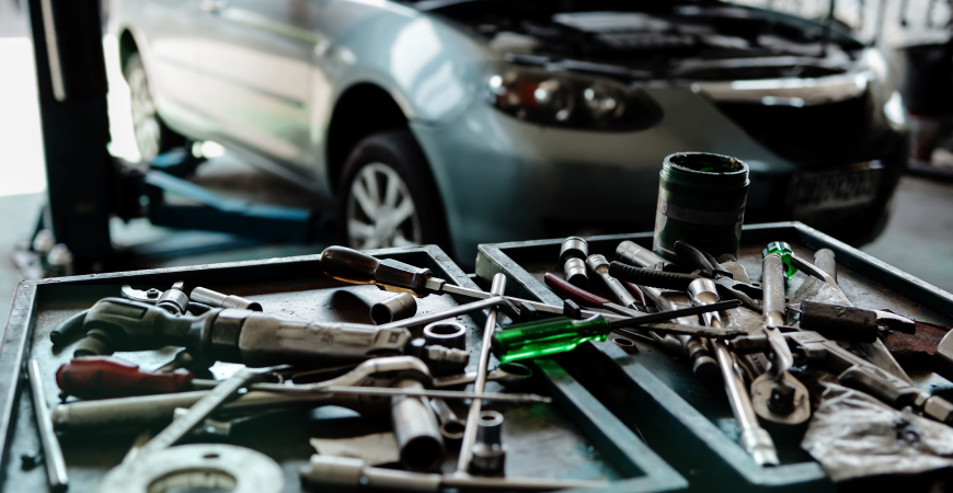 La imagen muestra una mesa de trabajo en un taller mecánico con varias herramientas esparcidas, incluyendo destornilladores, llaves inglesas y alicates. Al fondo, se puede ver un automóvil con el capó abierto, lo que sugiere que está siendo reparado o revisado. La escena es interesante y relevante porque ilustra el entorno típico de un taller de reparación de automóviles, destacando la variedad de herramientas utilizadas por los mecánicos y la atención que se presta a los vehículos en mantenimiento.