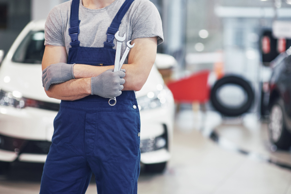 Un mecánico con guantes grises y un mono azul sostiene dos llaves en un taller de automóviles.