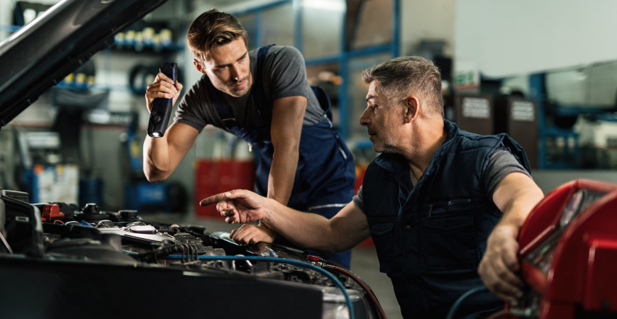 Dos mecánicos en un taller, uno sosteniendo una linterna y el otro señalando el motor de un automóvil.