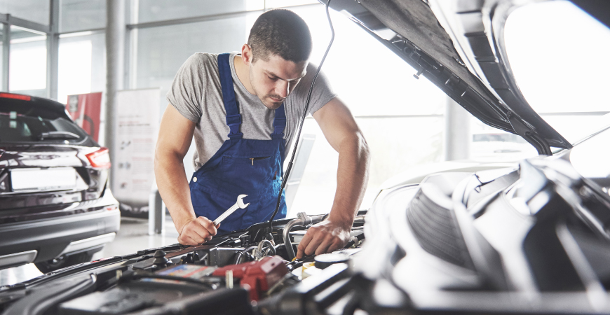 Una persona con un overol azul trabajando en el motor de un automóvil. La persona sostiene una llave inglesa y parece estar realizando algún tipo de reparación o mantenimiento. El automóvil tiene el capó levantado, y se pueden ver varias partes del motor. En el fondo, hay otro automóvil estacionado y algunas estructuras de un taller mecánico.