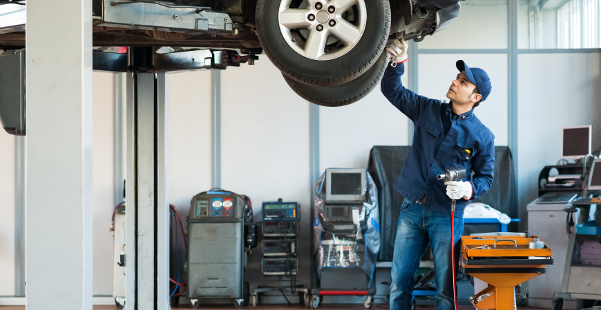 Esa imagen muestra un taller mecánico. En el centro, un mecánico está trabajando en un coche levantado, usando herramientas para arreglar la parte inferior. Varias máquinas, equipos de diagnóstico y una mesa con herramientas se encuentran al fondo, creando el típico entorno laboral de un taller.