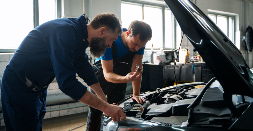 Dos personas trabajan en el motor de un automóvil en un taller mecánico, usando herramientas y discutiendo la reparación del vehículo.