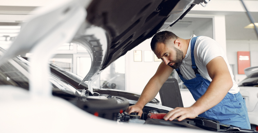 Un mecánico con un mono azul y una camiseta blanca está trabajando en el motor de un automóvil con el capó levantado en un taller mecánico.