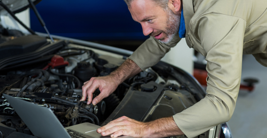 Un mecánico revisando el motor de un automóvil mientras consulta una computadora portátil.